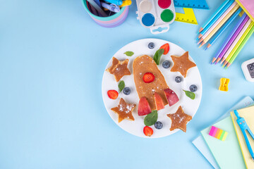 Back to school, school kids breakfast with creative shaped pancakes and fruit berries, on light blue background with school supplies, top view copy space. Kids hands eating pancakes