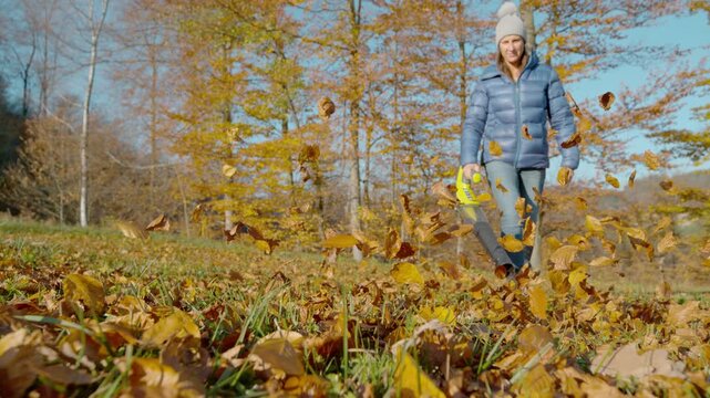SLOW MOTION: A woman operates a leaf blower amidst a colourful autumnal setting. Young lady clearing her garden lawn full of fallen autumn leaves from a nearby forest. Gardening chores in fall season.