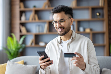 Close-up photo of a young Indian man sitting at home looking at the phone screen, holding a credit card