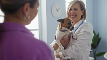 A mature blonde woman veterinarian in a clinic room indoors holding a small dog while another woman stands in the foreground, emphasizing the caring pet health environment.
