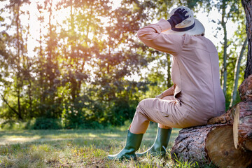 Woman gardener in a straw hat and garden uniform sitting on wooden tree logs and resting. Lush vegetation and trees in garden background. Sunny autumn day.