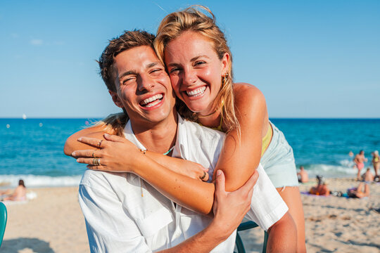 Close up portrait of a young Caucasian tourist couple hugging and looking at the camera on the beach while enjoying their summer vacation on the coast. Two people smiling together at a beach bar