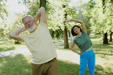 Senior silver-haired man and brunette woman dressed in cotton sportswear doing back exercise in park