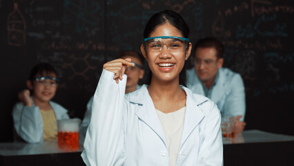 Girl looking at camera with arm folded while people doing experiment at laboratory. Cute student standing blackboard with chemical theory with blurring background at STEM science class. Edification.