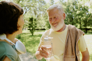 Gray-haired man holding bottle of water and smiling to his brunette wife after run in park