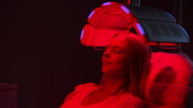 Closeup face of blonde female with closed eyes having LED red light hair photodynamic therapy during hair treatment in clinic. Young woman patient having non-invasive type of phototherapy, slow motion