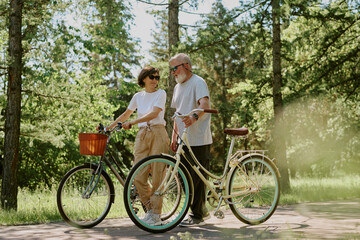 Modern senior couple having bicycle walk in local park