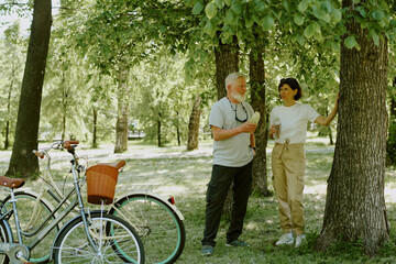 Senior couple resting and drinking water after ride while they standing between trees in park and chatting