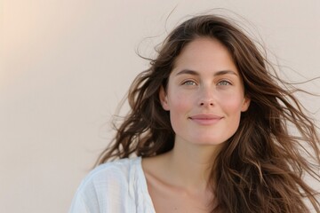 Naklejka premium Portrait of a smiling woman with long brown windblown hair, wearing a white top against a neutral background.