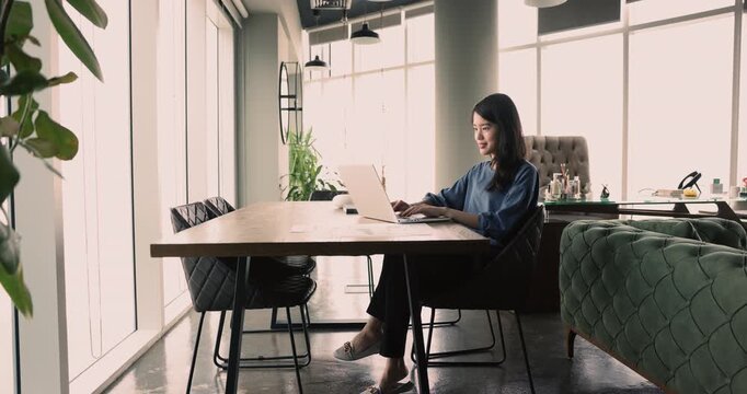 In modern office room young Asian woman working on-line on laptop, checks her email inbox for new business messages, responding to e-mails, correspond to clients, consulting remotely. Telecommuting