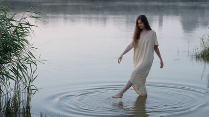 Fototapeta premium A dreamy girl or young woman in a simple light dress draws circles in the water with her feet in the middle of a lake or river