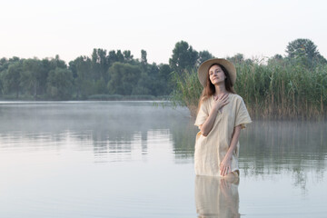A dreamy girl or young woman in a hat and a simple dress stands in a lake amidst the mist, smiling and touching herself with her hands