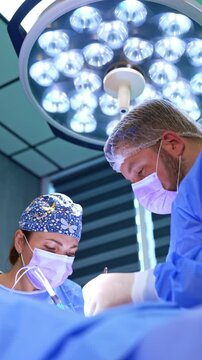 Surgeons use modern electric tool and traditional metal instrument at surgery. Cooperating doctors work under the round lamp in modern surgery room. Vertical video.
