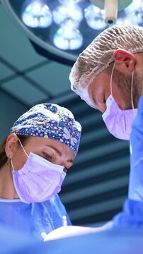 Concentrated faces of two doctors leaned over the patient. Diverse surgeons perform operation on thyroid. Low angle view. Vertical video.