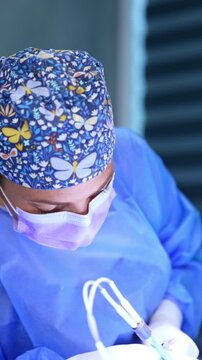 Female surgery professional wearing a cap with flowers and butterflies performs operation. Doctor applies modern electric surgical device in her work. Vertical video.