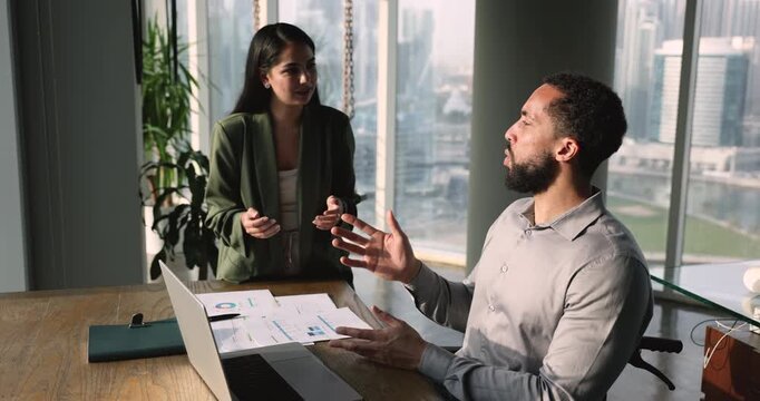 Female employee lead conversation with male colleague with disability, engaged in dialogue seated in wheelchair in modern downtown office. Opinion and ideas exchange, teamwork, collaborative project