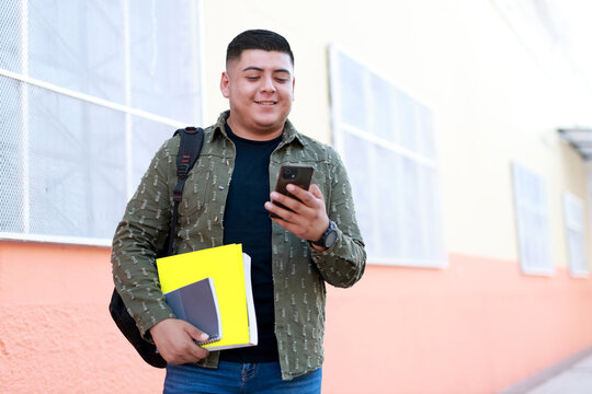 young latin american student holding books and smartphone outside the university