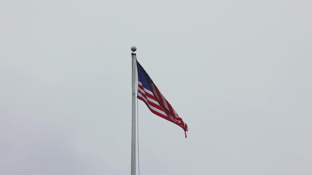 The U.S. flag flutters in the wind on a flagpole near the Dulles Airport terminal, with the cloudy sky as a backdrop, adding a patriotic tone to the scene.