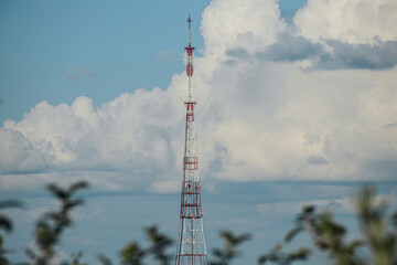 Obraz premium A tall metal radio tower, painted in red and white, stands prominently against a backdrop of a partly cloudy sky. The tower is surrounded by a landscape of trees and industrial structures