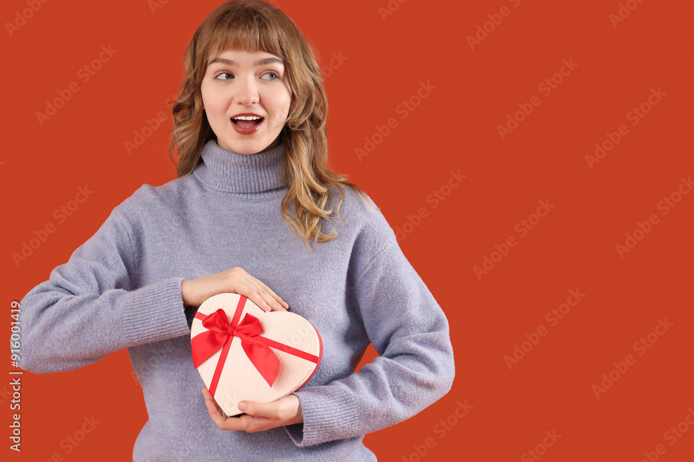 Poster Beautiful young shocked woman with gift box for Valentine's Day on orange background