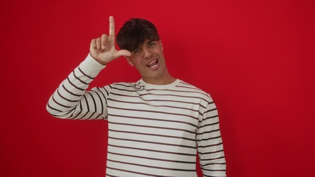 Loser gesture by young hispanic man wearing stripes t-shirt standing over isolated red background, mocking and insulting people with forehead fingers