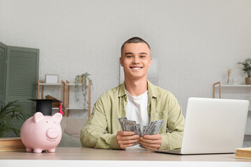 Male student with laptop at table with piggy bank and money in room. Concept of savings for...