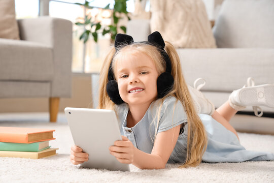 Cute little girl in headphones listening to audiobook with modern tablet computer while lying on carpet in living room