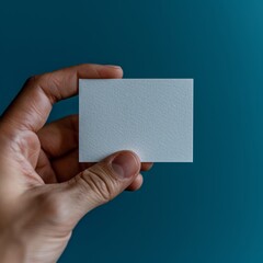 Person holding blank white business card against blue background. Card held between thumb and forefinger, in focus. Background slightly out of focus, solid blue color. Copy space.