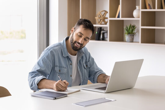 Positive Hispanic entrepreneur man using laptop for working in office, writing notes, smiling, talking on video call. Company manager, businessman watching professional learning webinar