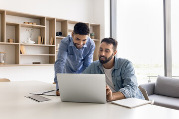 Two positive young diverse business professional men working at laptop together, discussing online project, smiling, looking at screen, sitting and standing at workplace table in co-working space