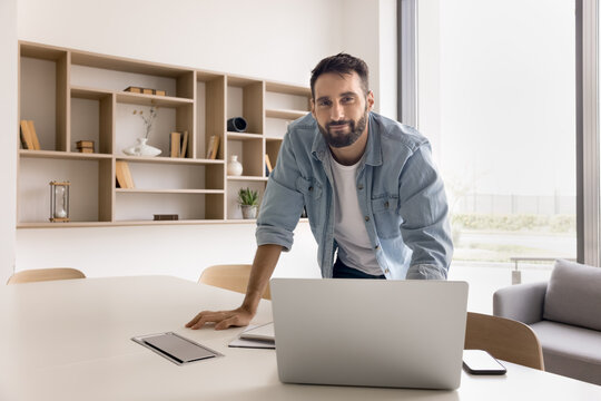 Positive confident Latin businessman, professional developer, entrepreneur, company owner workplace portrait. Handsome businessman leaning at table with laptop in office, looking at camera, smiling