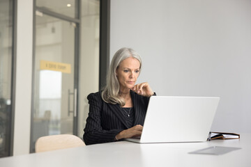 Focused elder company executive woman in suit working at laptop in meeting room alone, sitting at...