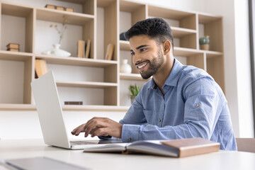 Happy handsome young Arab businessman typing on laptop at workplace table, chatting with client, enjoying online work communication, using professional application, smiling