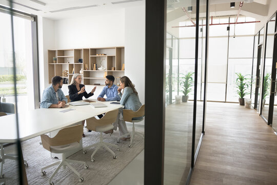 Diverse team of managers meeting for communication in office collaboration room behind glass wall, discussing startup at large table in modern workspace interior. Copy space with hall view