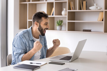 Naklejka premium Happy businessman excited with good news making winner hands at laptop, shouting yes, laughing, enjoying job success, work achievement, winning price, sitting at office workplace