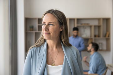 Thoughtful middle aged manager woman in casual standing in office workspace, looking away, thinking on business project, successful startup management with team talking in background