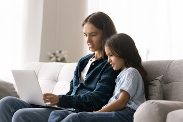 Positive focused young mom and daughter kid watching movie on laptop at home, using computer for communication, studying online application for school task, sitting with gadget on couch