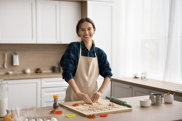 Happy attractive young baker girl in cooking apron preparing cookies for family holiday, cutting dough for biscuits, standing at table with bakery ingredients, looking at camera, smiling for portrait