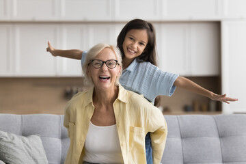 Active blonde grandma holding sweet beautiful kid girl from behind, looking at camera with toothy...