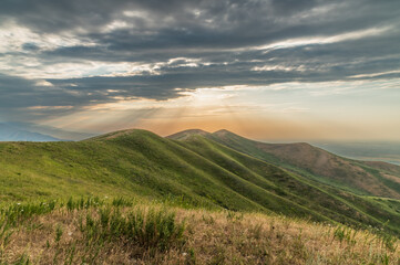 The sunset rays of the sun break through the clouds over green hills. Mystical sunset in the mountains. Stormy sky. Mountain landscapes of Kyrgyzstan.