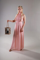 Pretty young blonde woman in pink evening dress holding vintage lantern in her hands on grey...