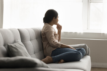 Thoughtful relaxed Indian woman resting seated on comfy couch in modern living room looking out...