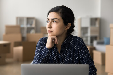 Young pensive woman working in warehouse, cardboard boxes on background, sit at desk with laptop...