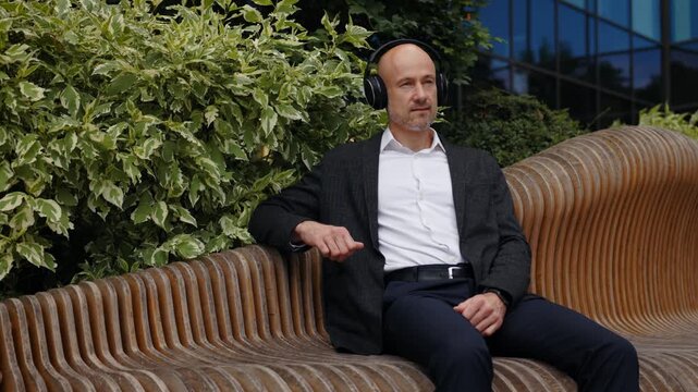 Bald businessman is sitting on bench with headphones on, listening to hard music, dancing slightly. Man is dressed in dark jacket and white shirt. In background, there is greenery