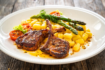 Lamb chops with bone, gnocchi and cooked green asparagus served on white plate on wooden table
