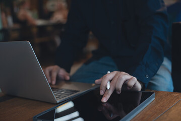 Man working on laptop computer and using digital tablet surfing the internet on wooden table at coffee shop