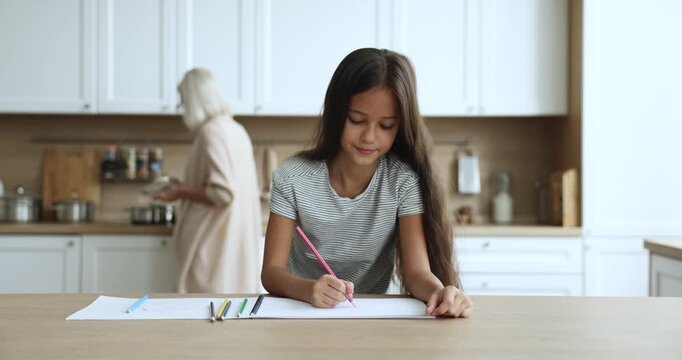 Focused preteen kid girl drawing in colorful pencils in paper album, sitting at kitchen table while blonde grandmother cooking lunch behind. Positive schoolkid doing school homework at home