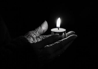 Black and white photography of an old woman's hand holding a candle, dark background with light coming from the flame. The photograph is in the style of a minimalist portrait.