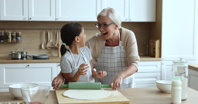 Cheerful baker grandmother and granddaughter girl in aprons having fun in kitchen, touching noses with floury hands, baking homemade pastry food, laughing, smiling