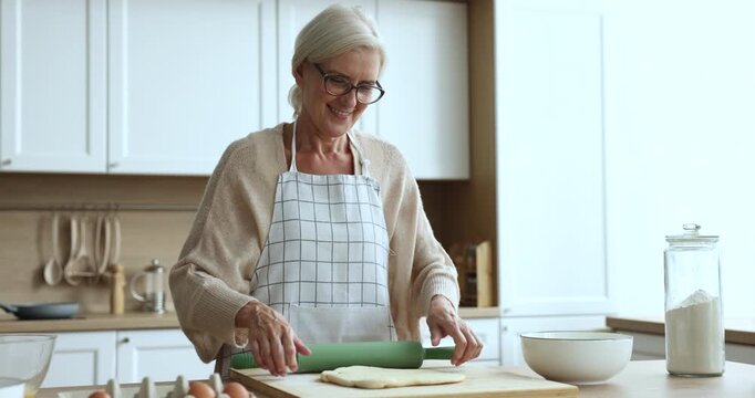 Happy blonde senior baker woman in kitchen apron rolling dough on table with bakery ingredients, preparing pastry, baking dessert, cake, pie, cookies, smiling, enjoying culinary hobby